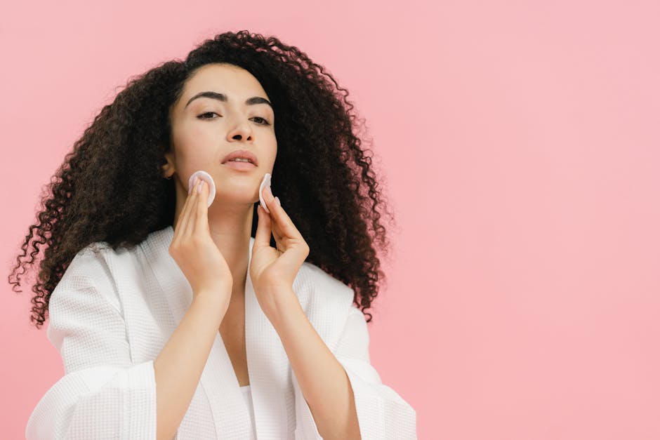 A young woman with curly hair using cotton pads for her skincare routine against a pink background
